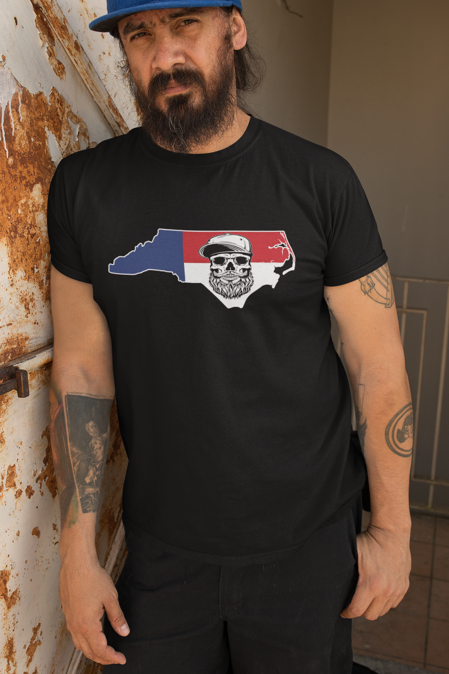 Man wearing a black t-shirt with a graphic design of a state map with a skull and crossed axes, standing against a rusty metal wall.