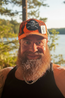 Man with a beard wearing an orange cap with a skull patch, standing outdoors by a body of water.