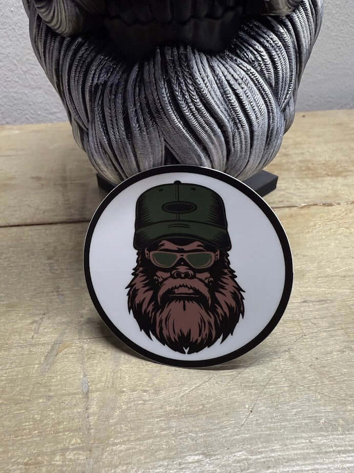 Bearded Bigfoot Yeti sticker featuring sunglasses and a cap on a wooden surface.