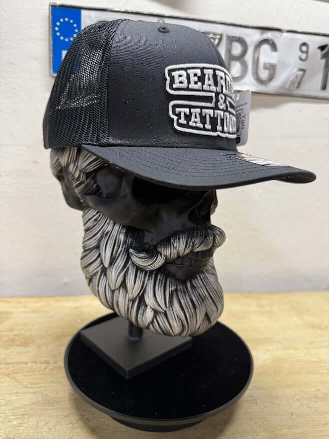 Bearded and tattooed trucker hat displayed on a skull bust, showcasing bold design and mesh back for a rugged look.